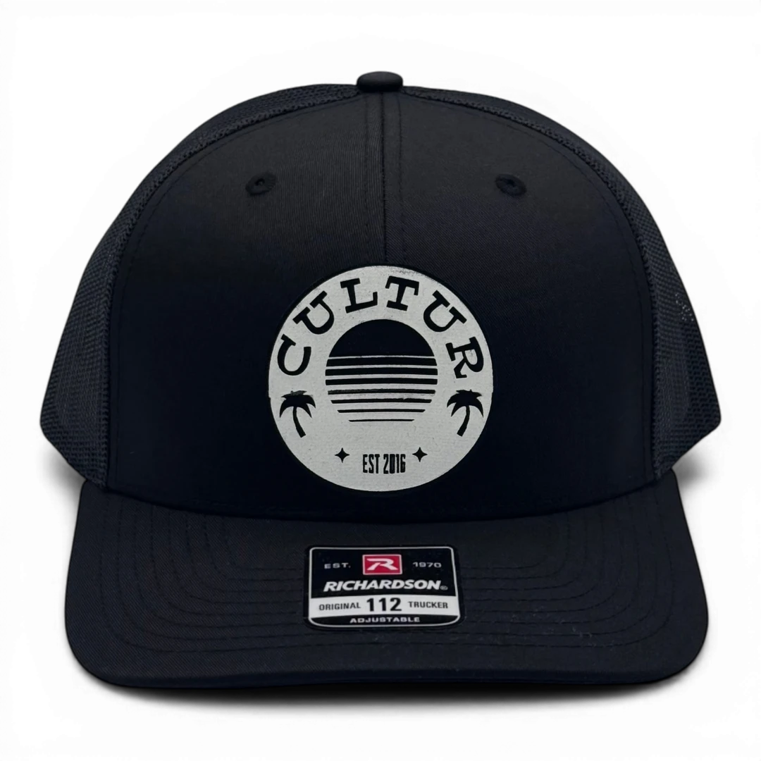 CULTUR black trucker hat with mesh back and circular patch featuring a sunset and palm trees.