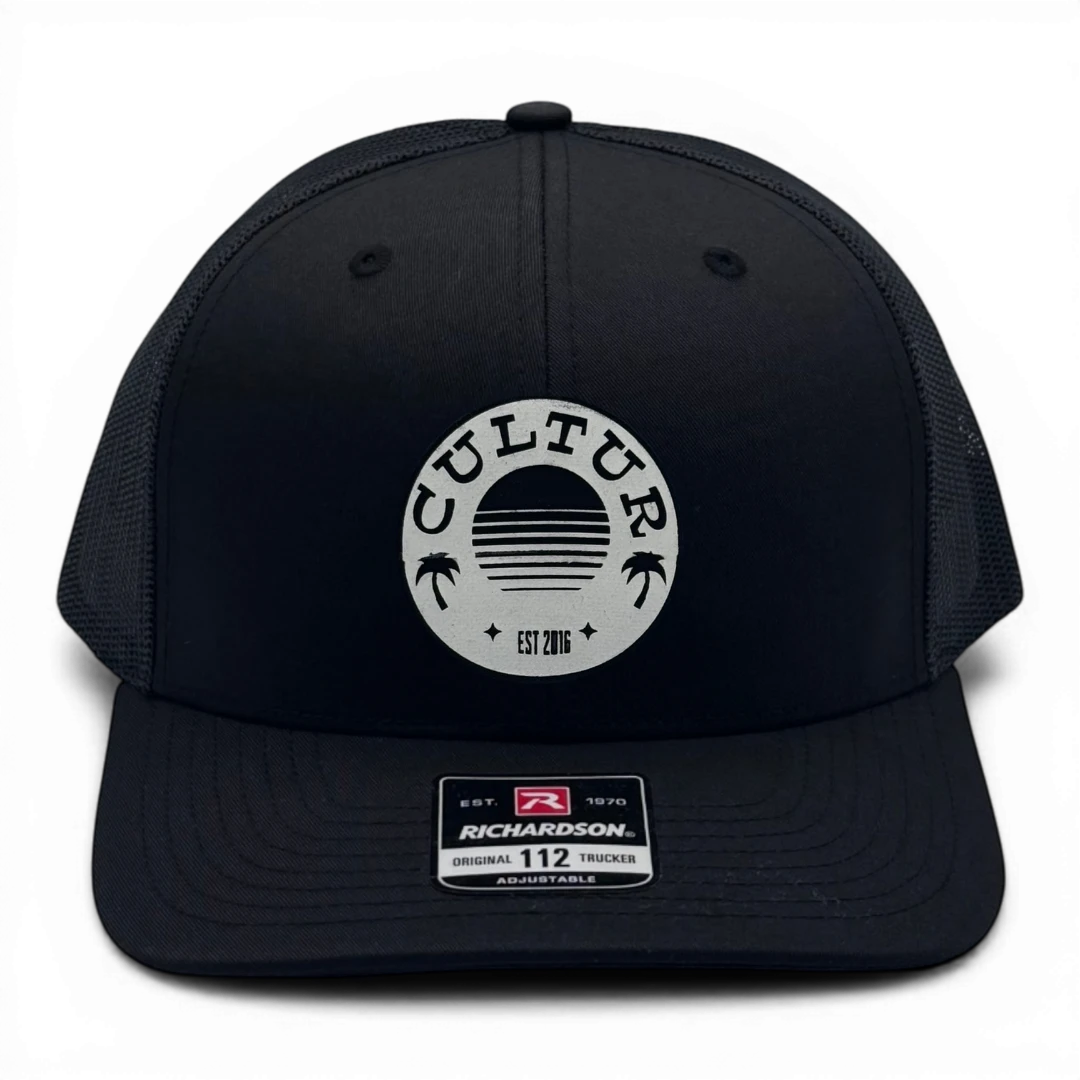 CULTUR black trucker hat with mesh back and circular logo featuring a sunset design with palm trees and "EST 2016".