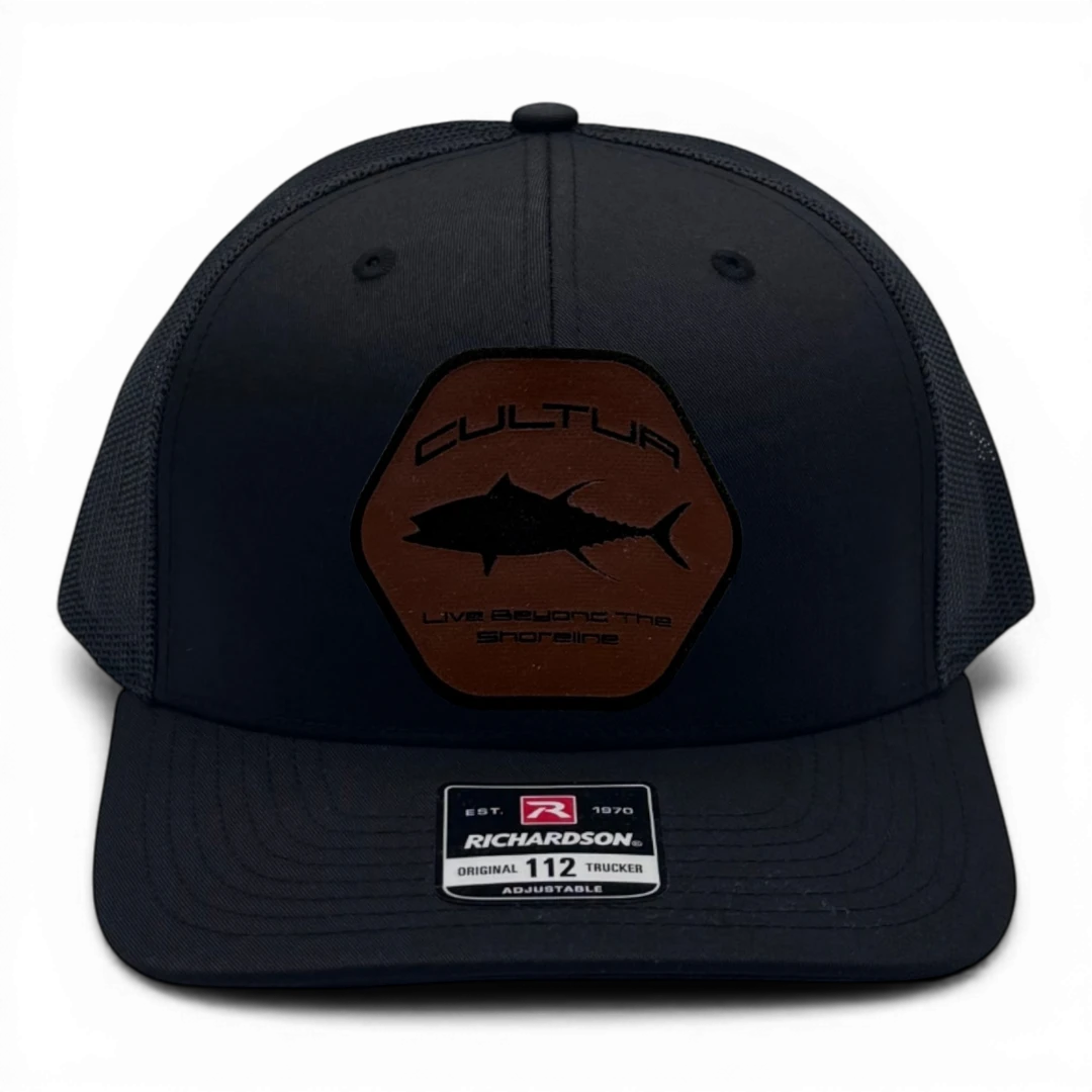 CULTUR black trucker hat with a hexagonal brown patch featuring a tuna silhouette and "Live Beyond The Shoreline" text.