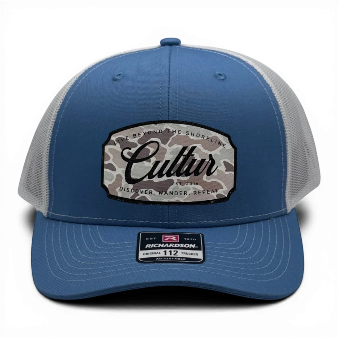 CULTUR blue trucker hat with white mesh back and a camouflage patch featuring the CULTUR logo.
