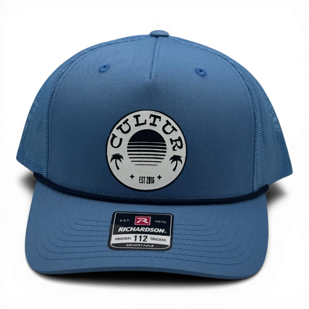 CULTUR blue trucker hat with mesh back and circular patch featuring palm trees and a sunset design.