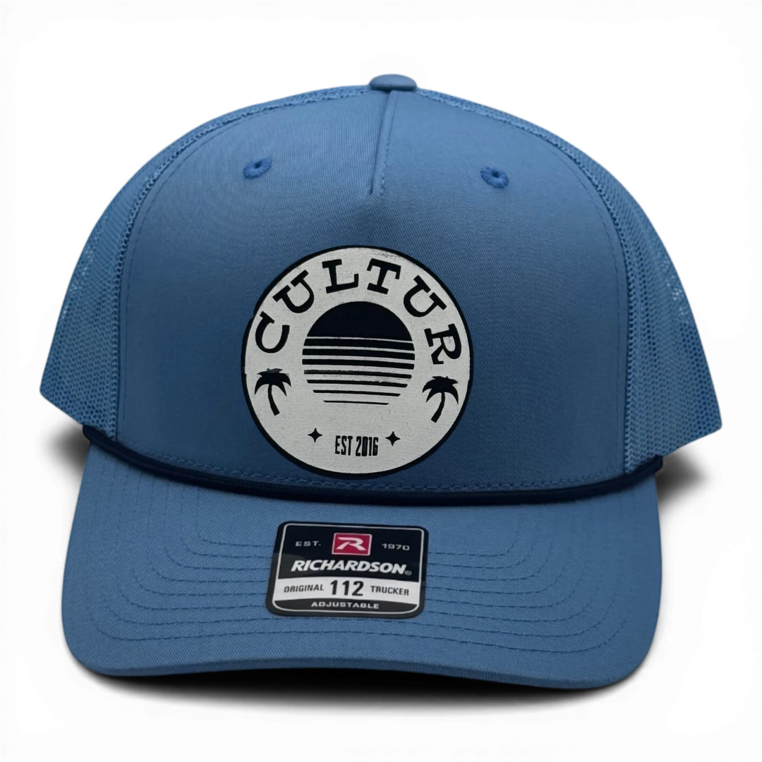 CULTUR blue trucker hat with mesh back and circular patch featuring palm trees and a sunset.
