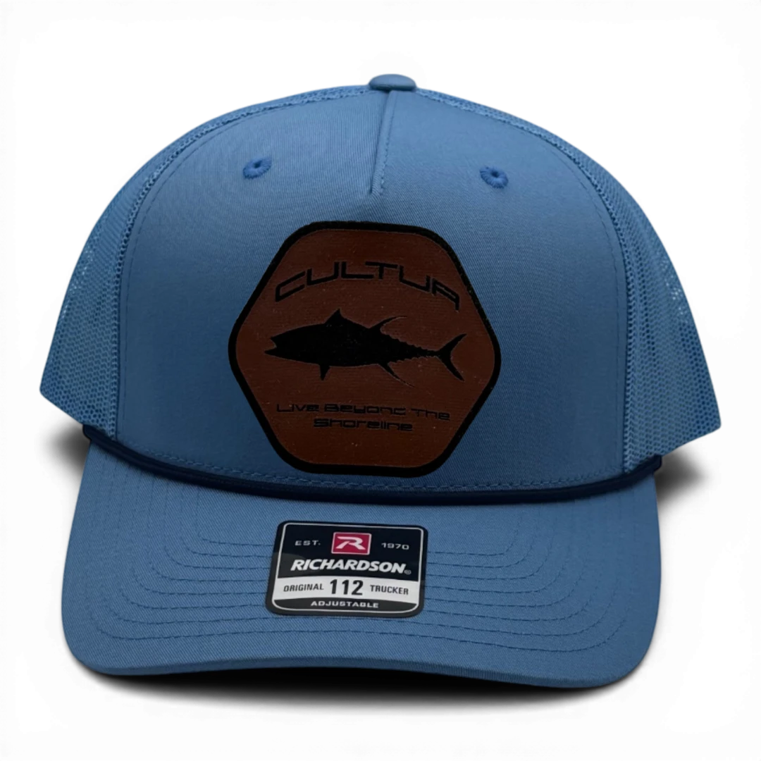CULTUR blue trucker hat with a hexagonal patch featuring a tuna silhouette and "Live Beyond The Shoreline" text.