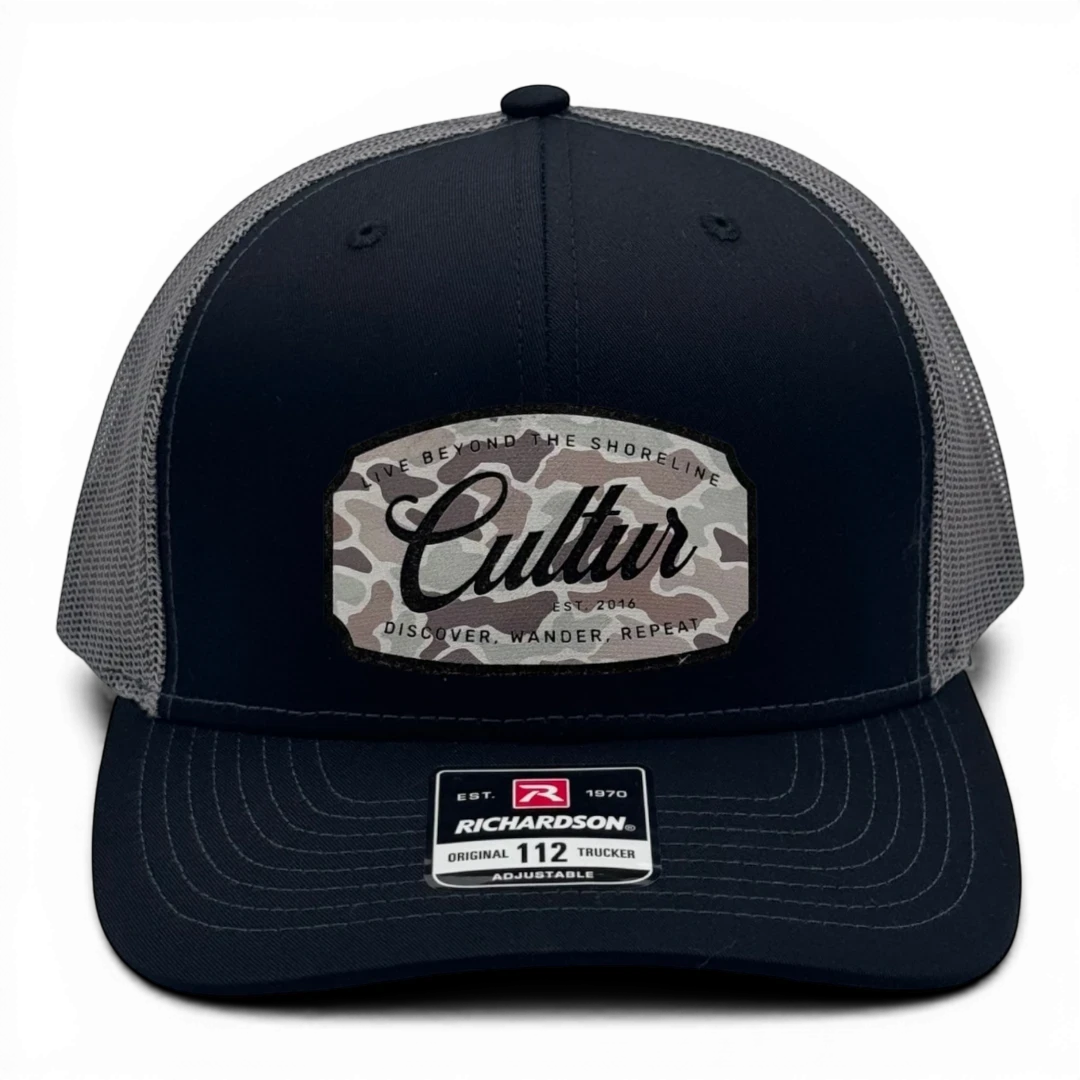 CULTUR black trucker hat with grey mesh back and a camouflage patch featuring the CULTUR logo and text.