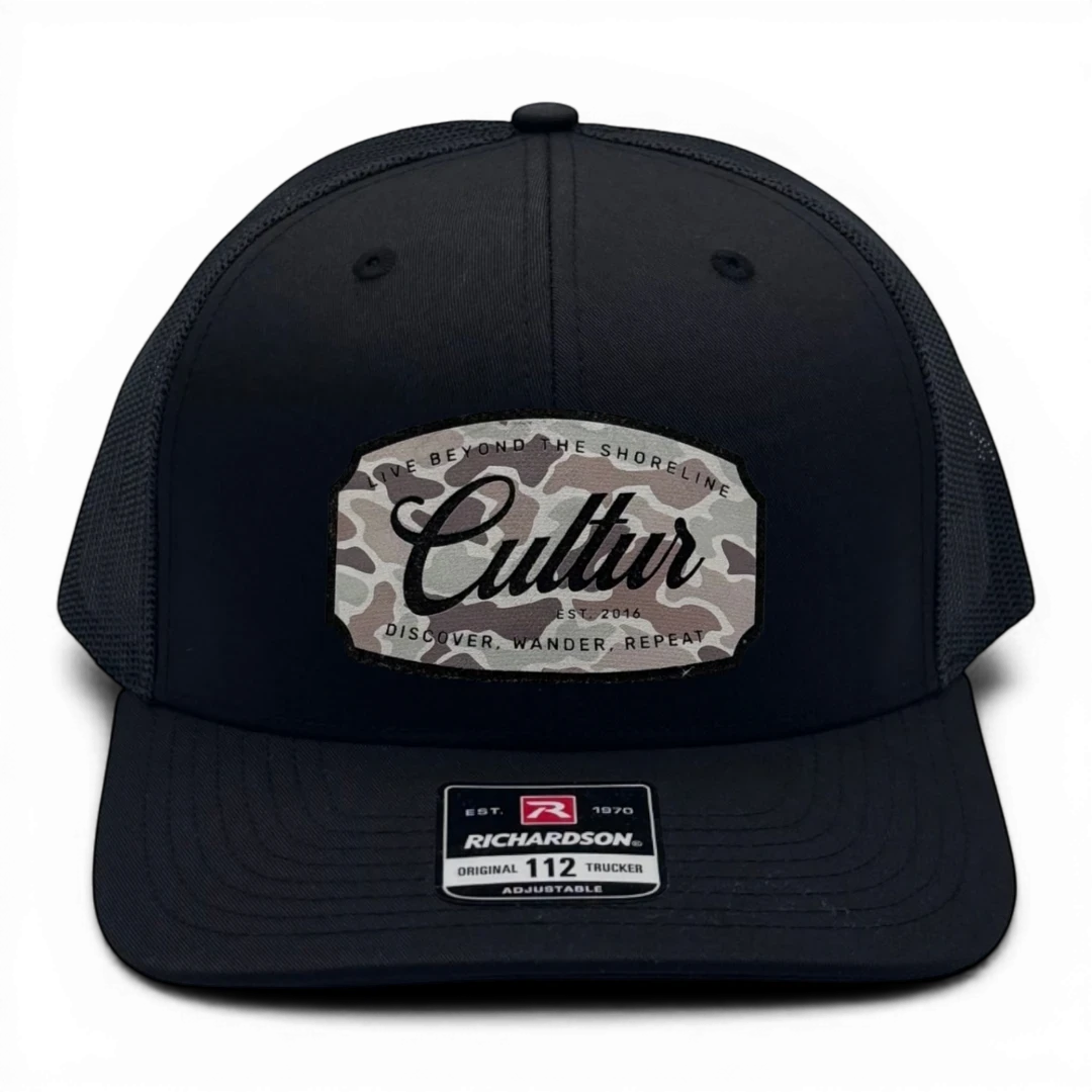 CULTUR black trucker hat with a camouflage patch featuring the CULTUR logo and text "LIVE BEYOND THE SHORELINE, DISCOVER, WANDER, REPEAT."