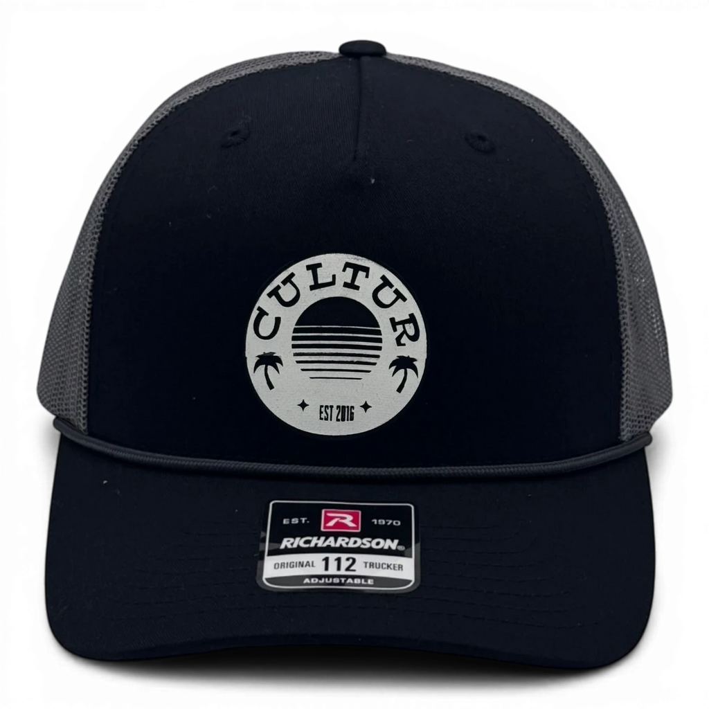 CULTUR black trucker hat with grey mesh back and circular CULTUR patch with palm trees and sun graphic.