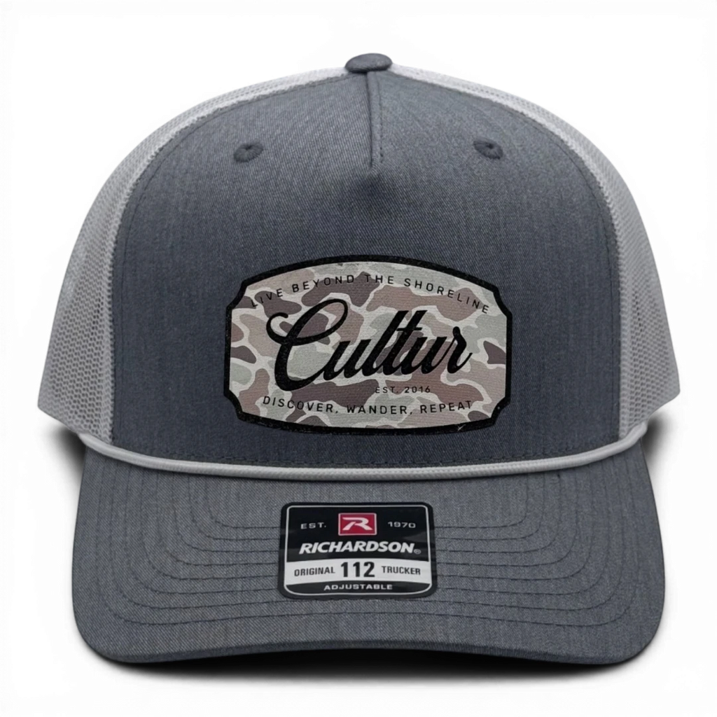 CULTUR grey trucker hat with white mesh back and a camouflage patch featuring the CULTUR logo.