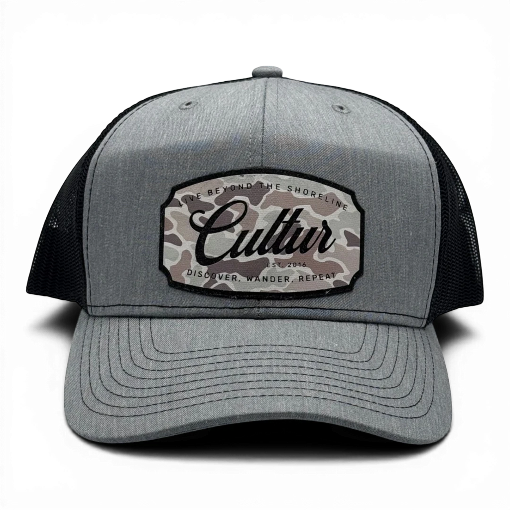 CULTUR grey trucker hat with black mesh back and a camouflage patch featuring the CULTUR logo.