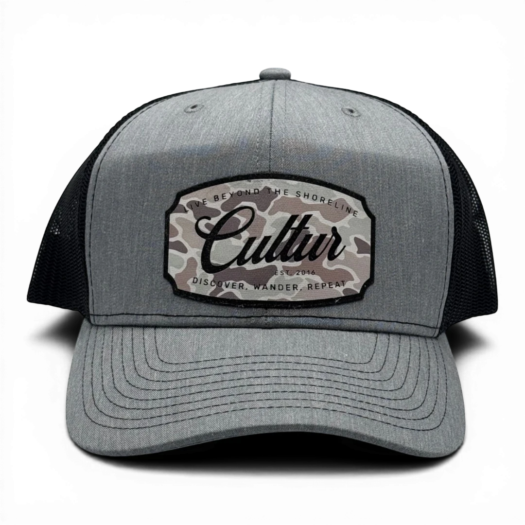 CULTUR grey trucker hat with black mesh back and a camouflage patch featuring the CULTUR logo.
