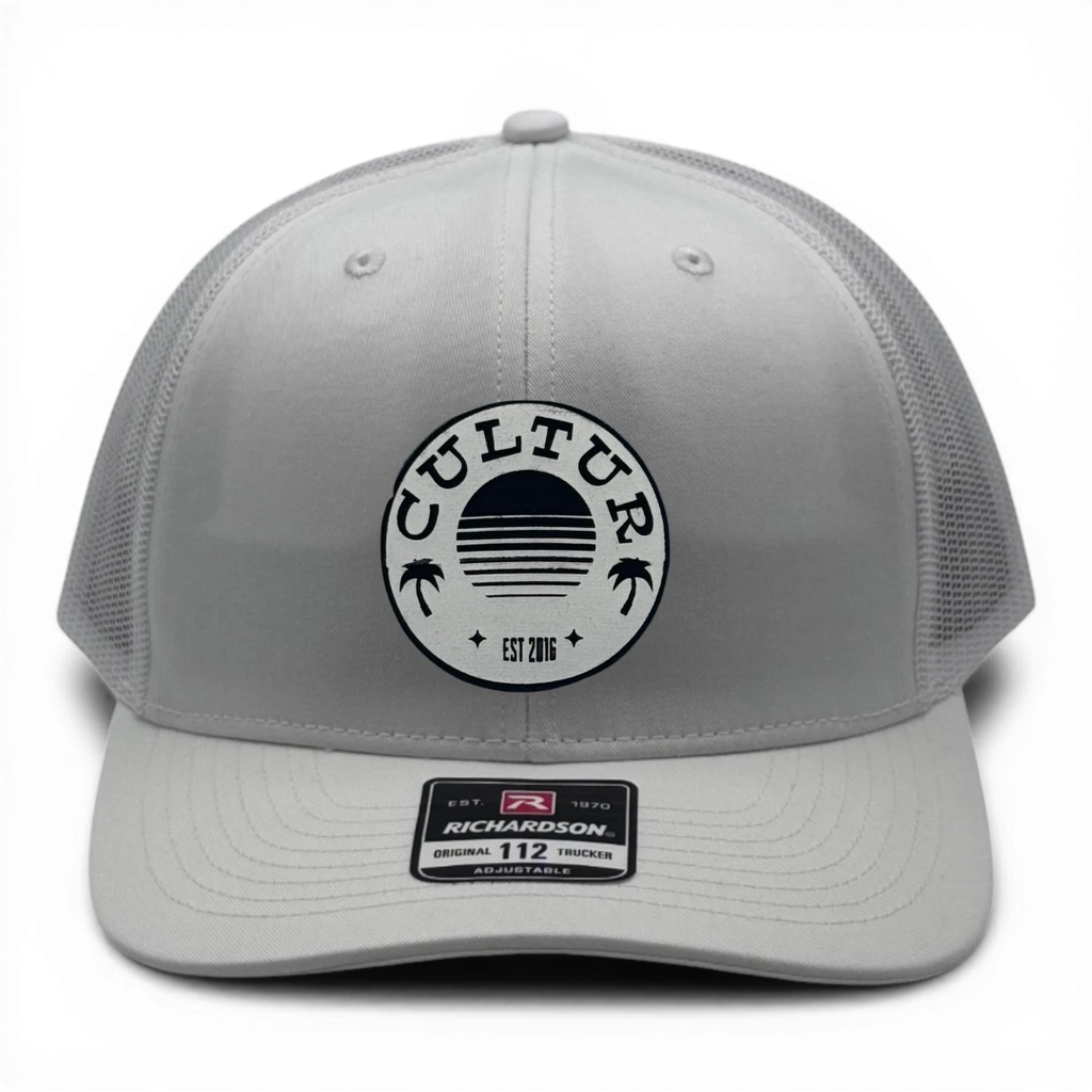 CULTUR white trucker hat with mesh back and circular logo featuring palm trees and a sunset.