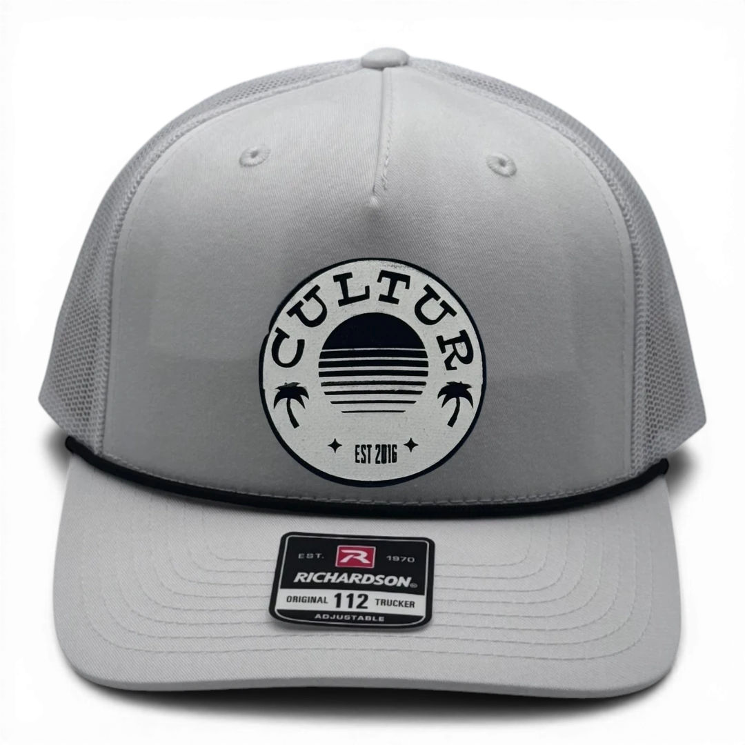 CULTUR white trucker hat with black rope and mesh back and circular patch featuring a sunset and palm trees.