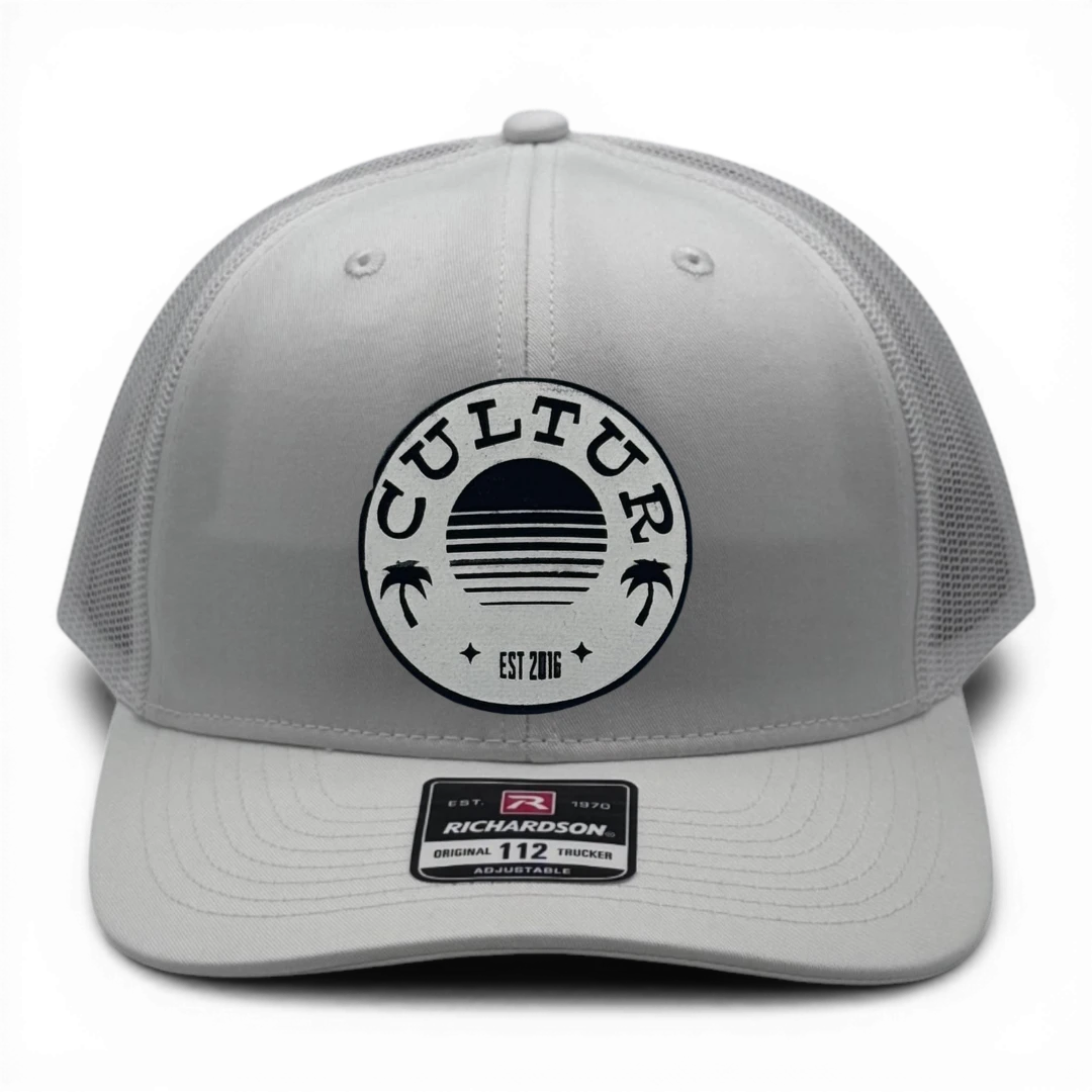 CULTUR white trucker hat with a circular patch featuring a sunset and palm trees, adjustable fit.